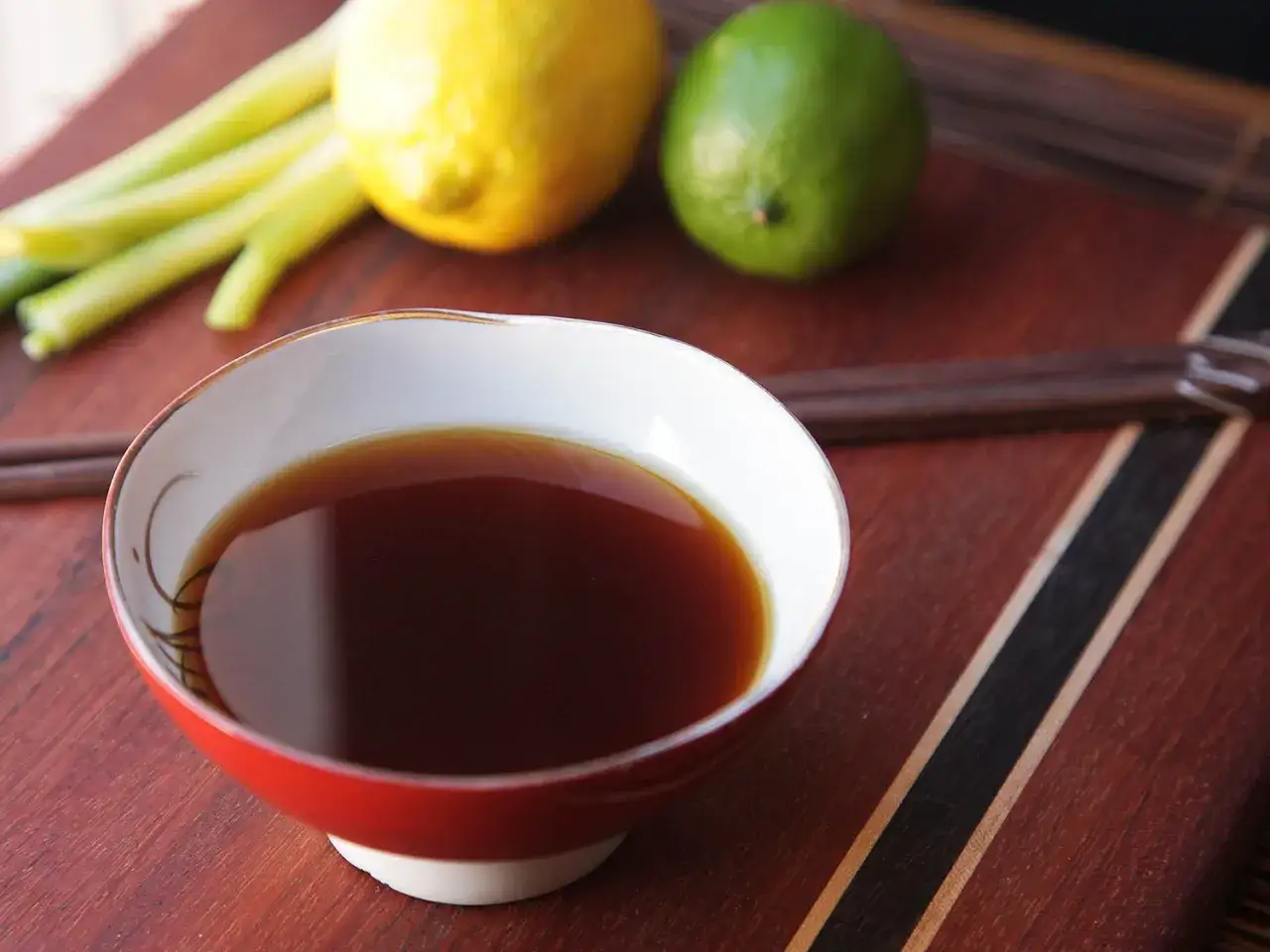 A bowl of dark liquid, possibly a ponzu sauce substitute, sits on a wooden board with a lemon, lime, and celery stalks.