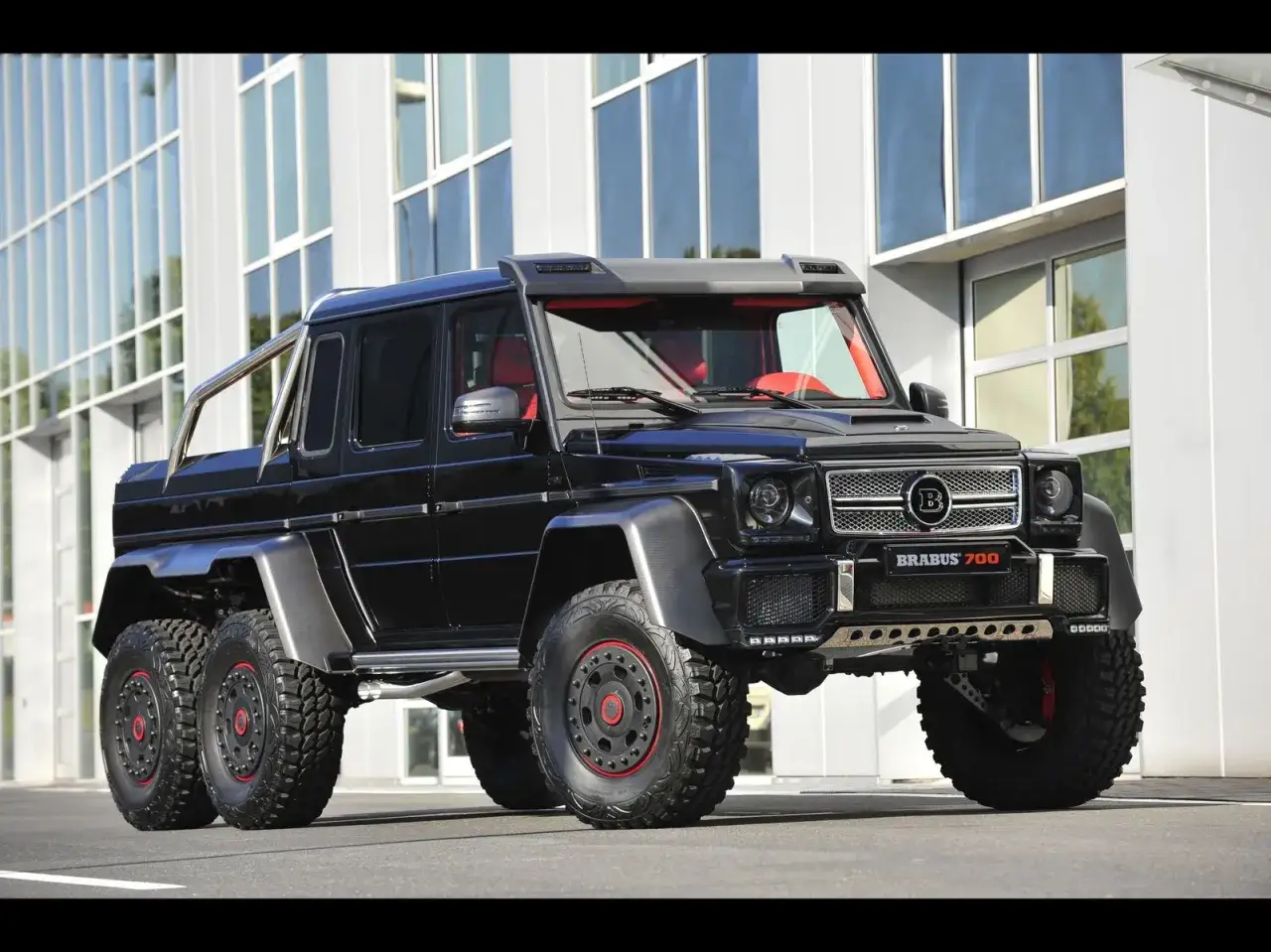 Czarny, sześciokołowy Brabus 700 z czerwonym wnętrzem, zaparkowany przed nowoczesnym budynkiem.