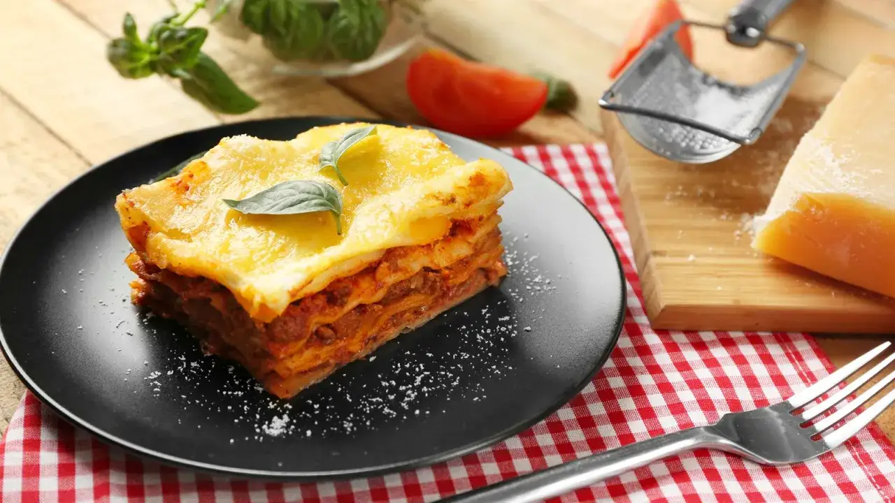 Ein St&uuml;ck Lasagne mit reichlich Bechamelsauce und K&auml;se &uuml;berbacken, garniert mit Basilikum. Daneben ein St&uuml;ck Parmesan und eine Gabel.