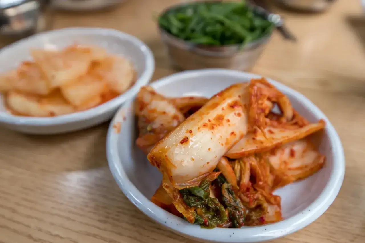 Kapusta kimchi co to jest? Pikantna, fermentowana kapusta pekińska w miseczce, obok innych koreańskich przystawek.