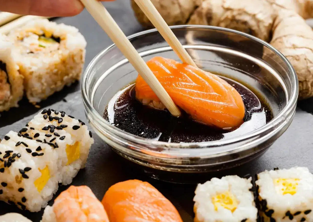 Paleczki zanurzają kawałek łososia w ciemnym sosie sojowym, obok sushi i imbiru.