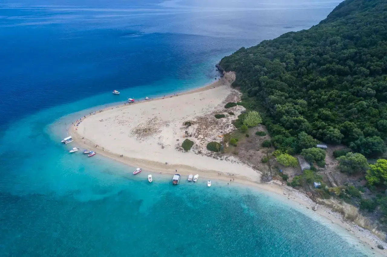 Plaża na wyspie greckiej Zakynthos, otoczona turkusowym morzem i zielonym lasem. Kilka łodzi zacumowanych przy brzegu, ludzie spacerują po piasku.