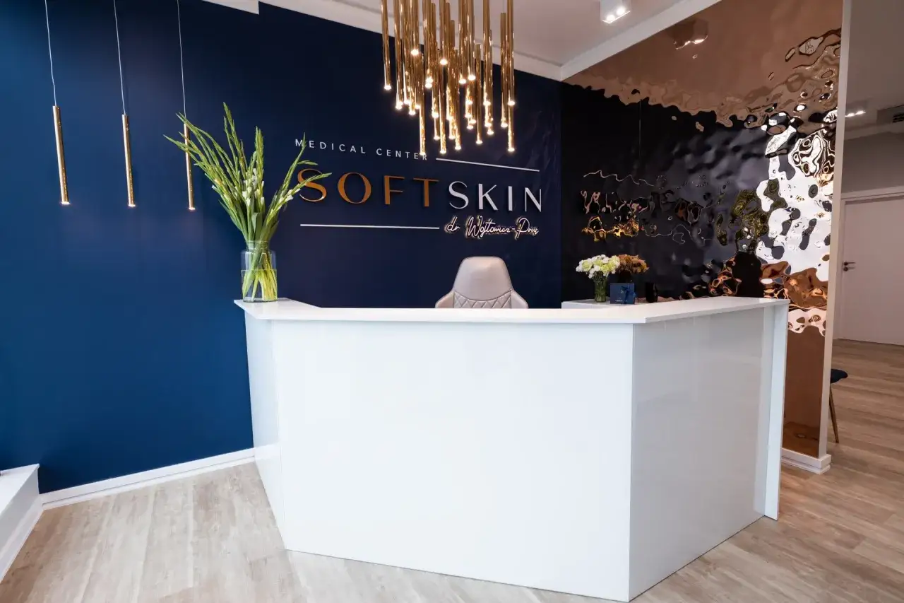 Nowoczesne wnętrze recepcji kliniki medycznej Soft Skin z białym lady, ciemnoniebieską ścianą i złotymi lampami.