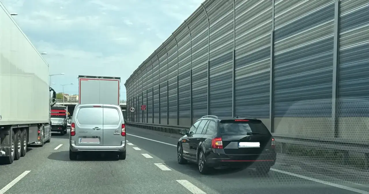 Samochody i ciężarówki na autostradzie, przygotowujące się do **jazdy na suwak**. Długie ściany dźwiękochłonne po prawej stronie.