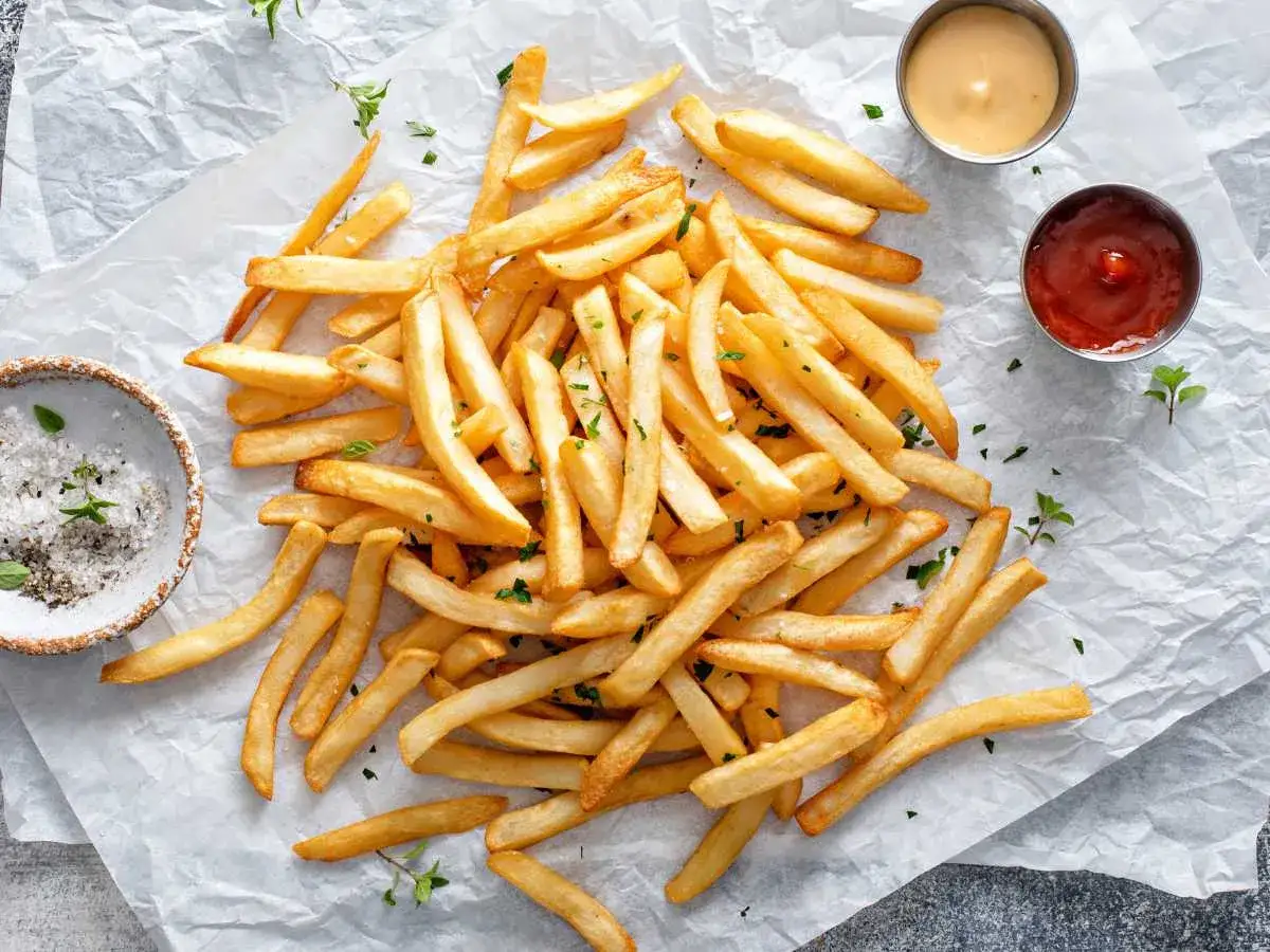 Montón de patatas fritas airfryer doradas y crujientes, espolvoreadas con perejil picado, junto a un cuenco de sal y dos salsas.