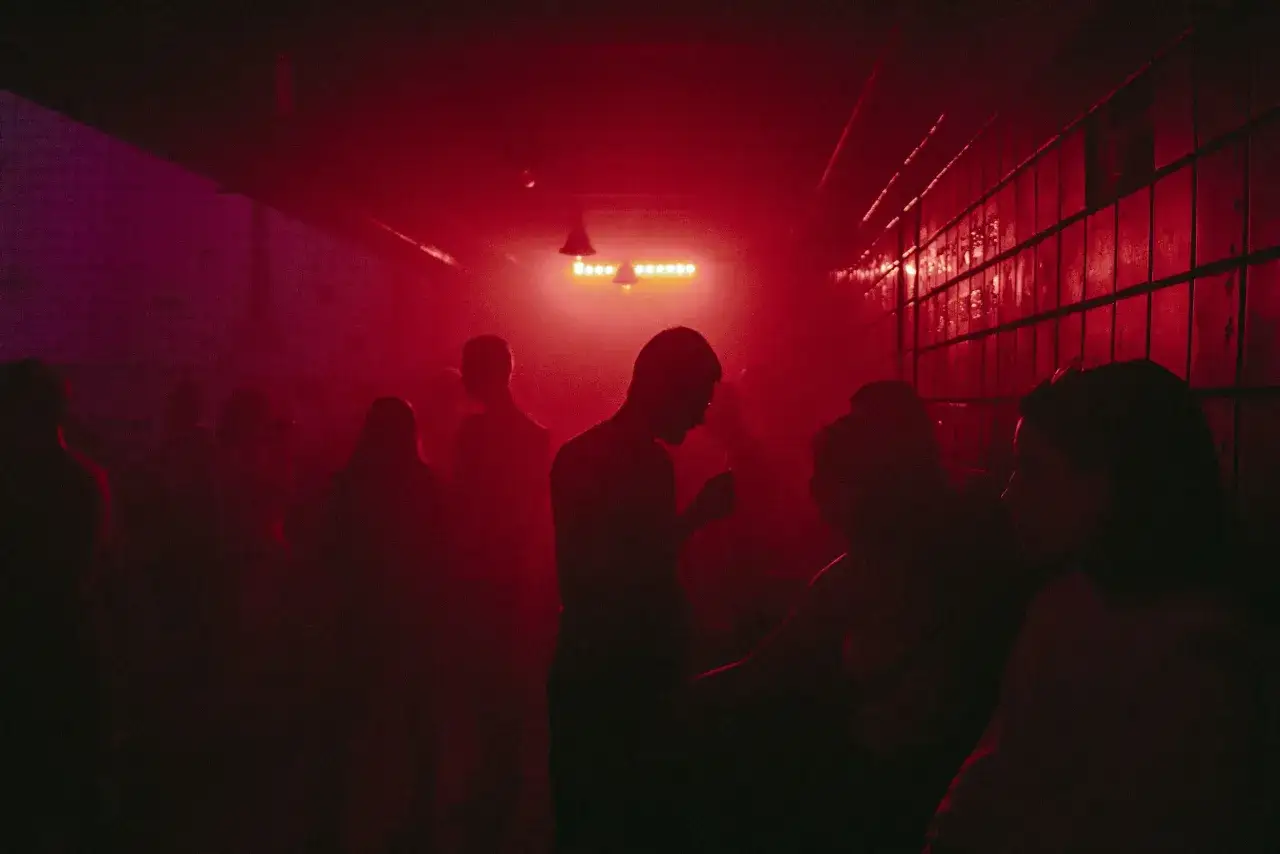 Silhouetten von Menschen tanzen im roten Licht einer Techno-Party. Nebel und pulsierende Lichter schaffen eine hypnotische Atmosphäre.