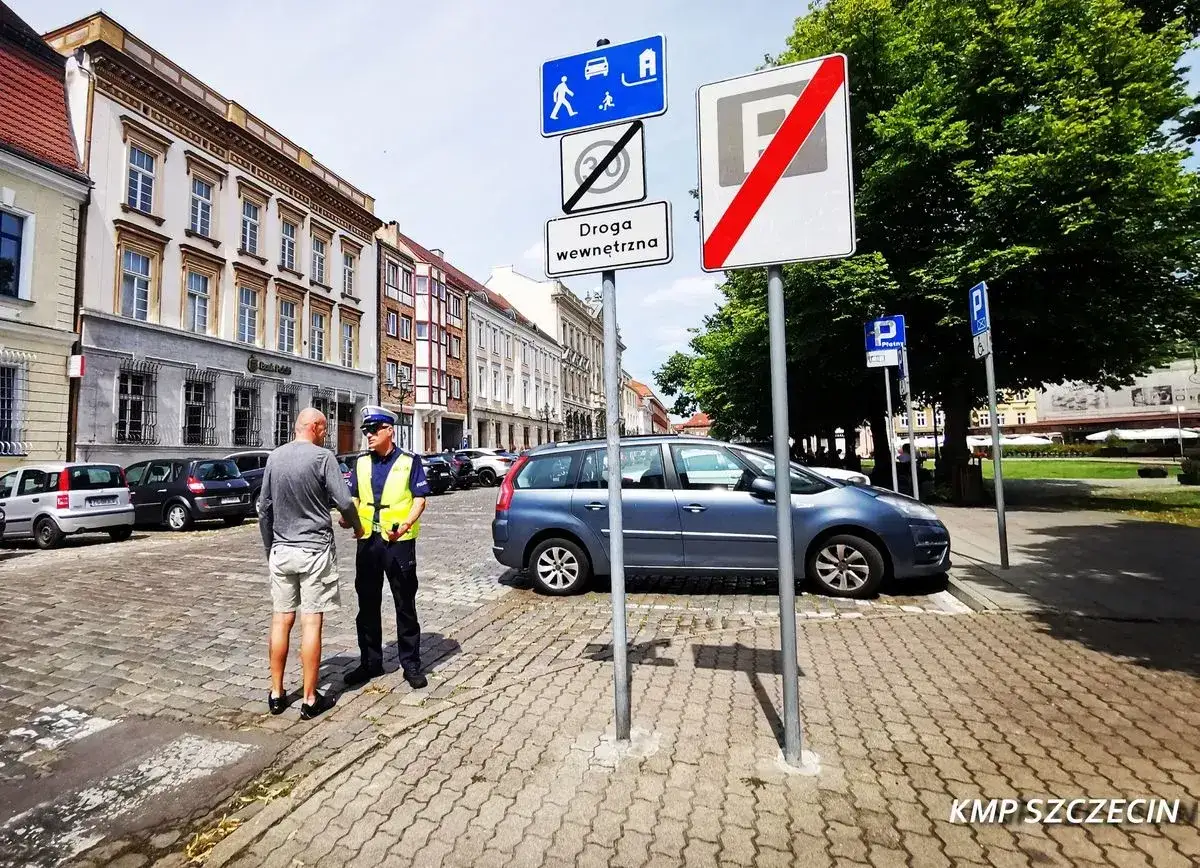 Policjant rozmawia z mężczyzną na brukowanej ulicy. Znaki drogowe wskazują koniec strefy zamieszkania i ograniczenie prędkości.