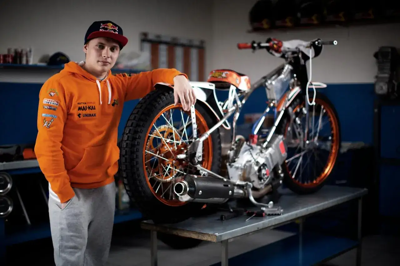 Młody zawodnik w pomarańczowej bluzie opiera się o swój motocykl żużlowy, gotowy do wyścigu.