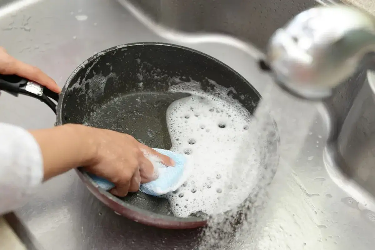 Eine Hand reinigt eine beschichtete Pfanne, die klebt, unter flie&szlig;endem Wasser mit einem Schwamm und Seifenschaum in der Sp&uuml;le.