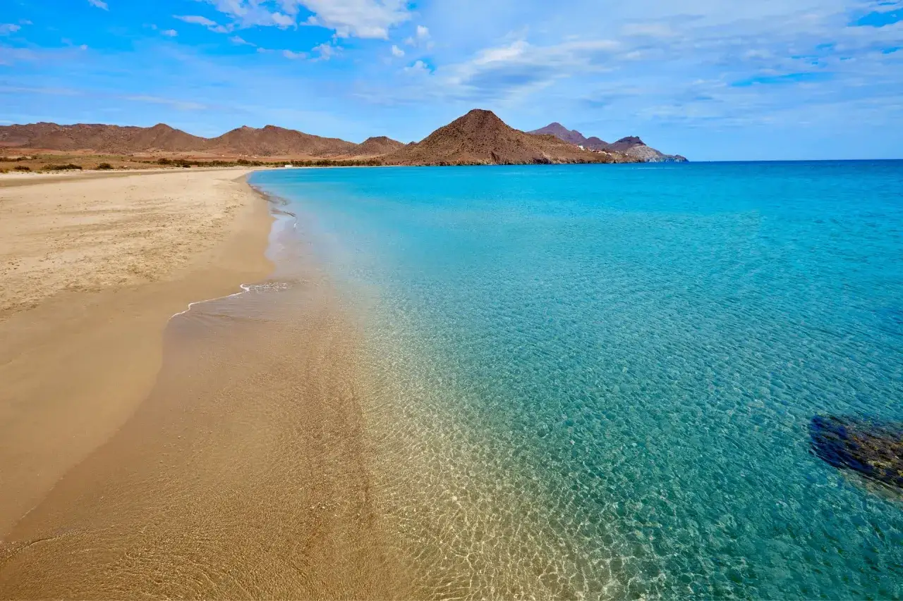 Arena dorada y aguas cristalinas en las playas bonitas de Cabo de Gata. Monta&ntilde;as &aacute;ridas al fondo bajo un cielo azul con nubes.