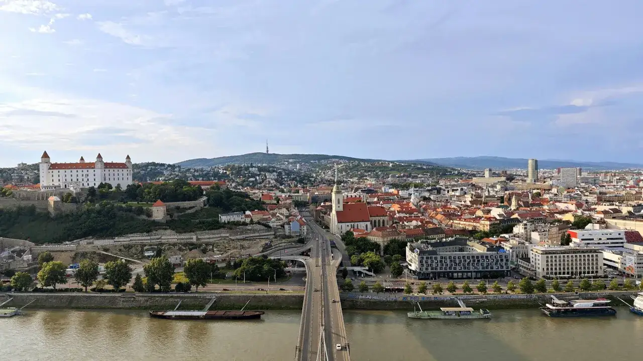 Widok na Dunaj i bratysławskie stare miasto z zamkiem na wzgórzu. Most łączy brzegi, a w oddali widać wzgórza.