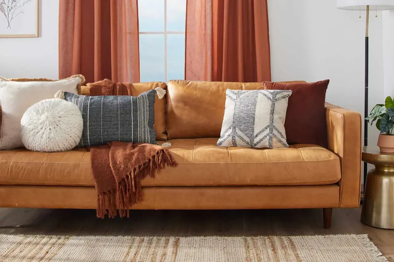 A warm, caramel-colored leather couch showcases a variety of plush couch materials: a chunky knit round pillow, a striped lumbar pillow with tassels, a textured throw blanket, and patterned square cushions.