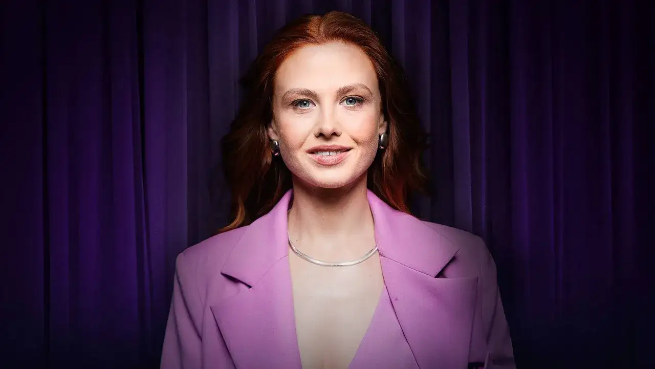 Anna Karczmarczyk w różowej marynarce, z rudymi włosami i niebieskimi oczami, uśmiecha się na tle fioletowej kurtyny. Zapowiada swoje filmy, seriale i programy.