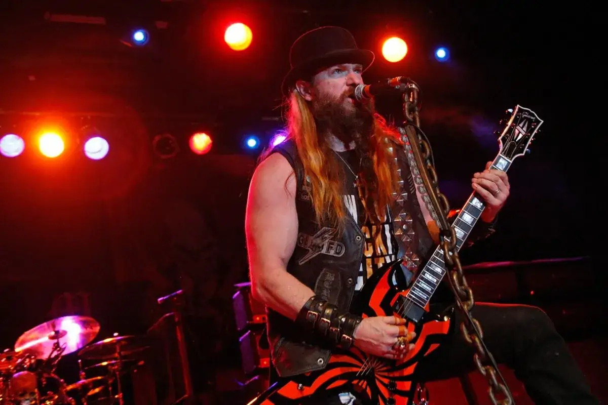 Zakk Wylde z zespołem na scenie, w czarnym kapeluszu i kamizelce, gra na gitarze.