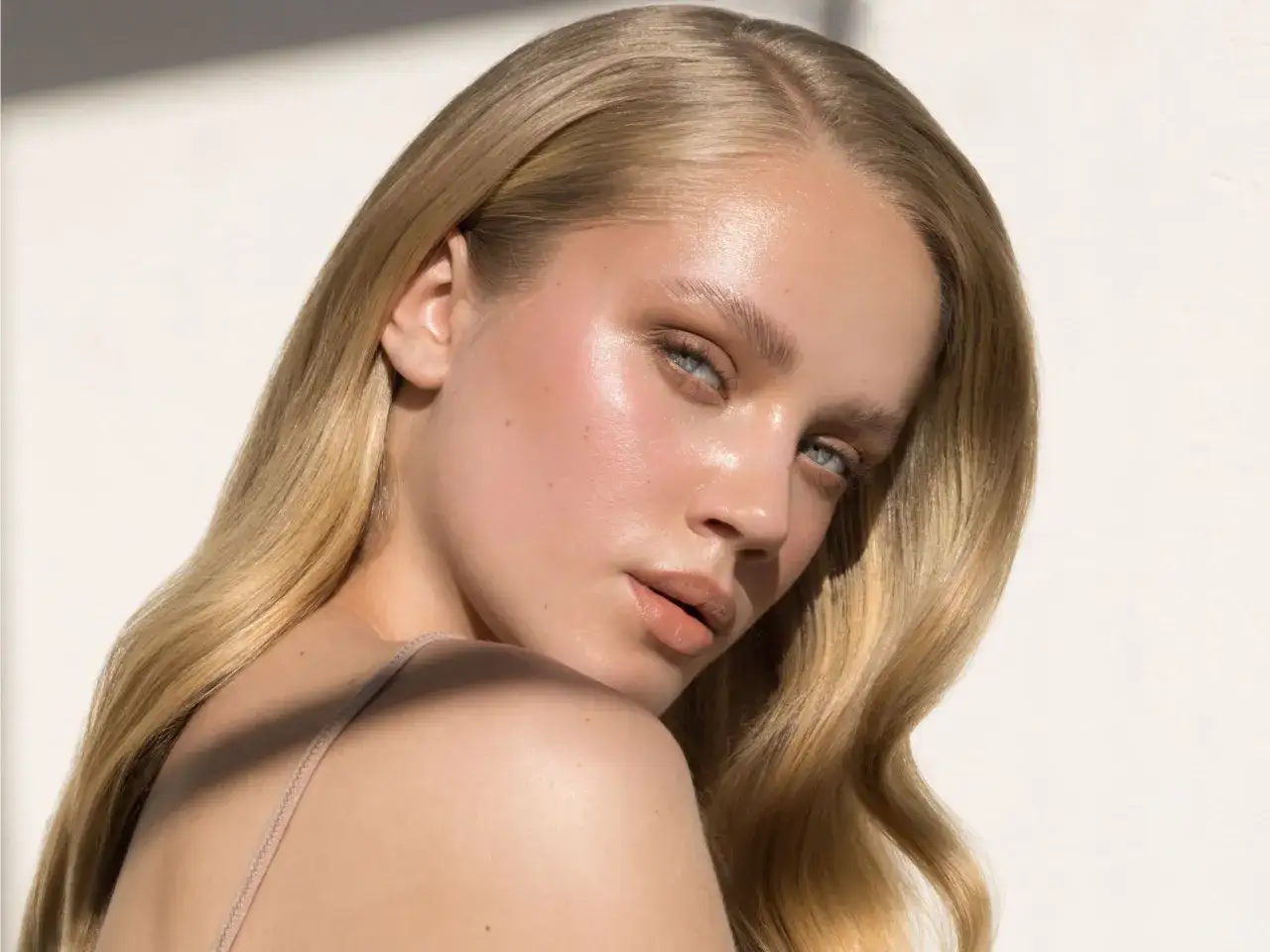 Rostro de mujer con piel luminosa y dewy, cabello rubio ondulado y ojos azules.