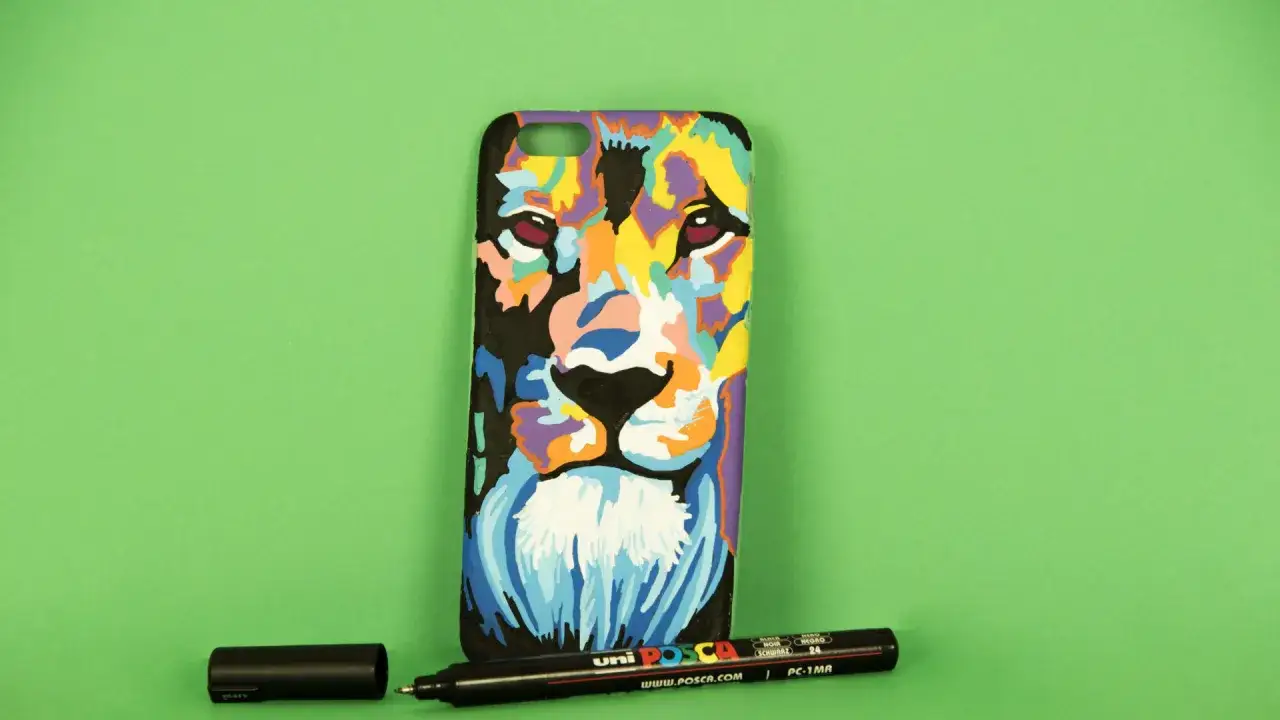 Coque de téléphone ornée d'un lion coloré, peinte avec des marqueurs Posca. Un vernis fixateur Posca est posé à côté.