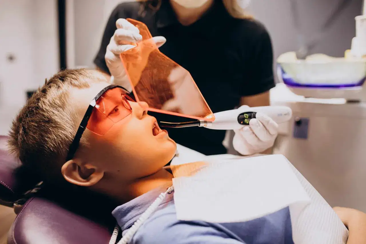 Chłopiec w okularach ochronnych przechodzi laserowe leczenie zębów. Dentysta używa urządzenia emitującego światło.