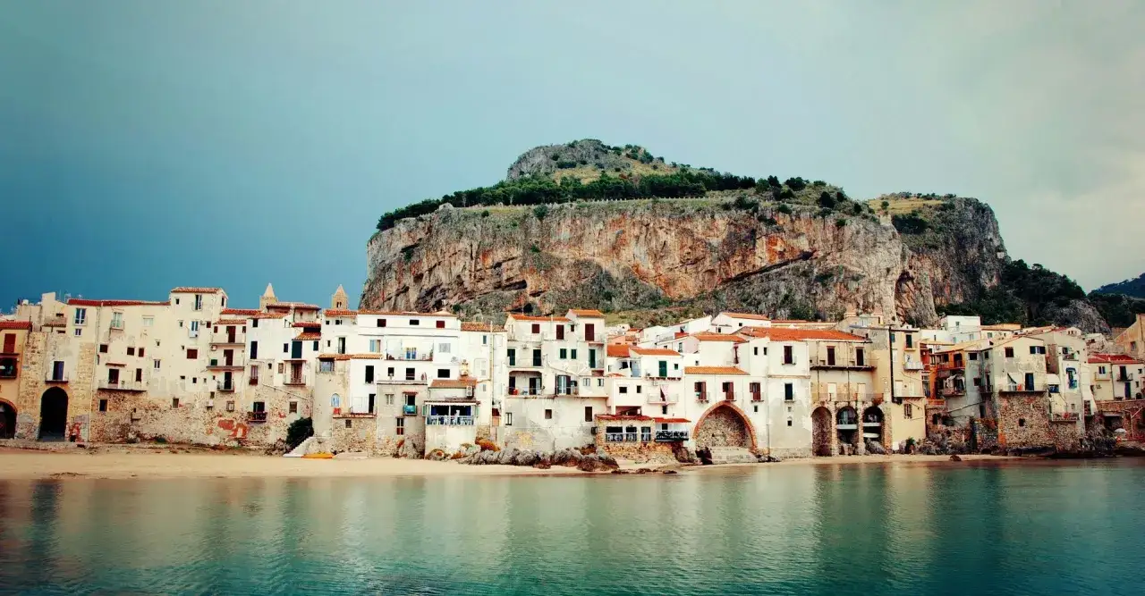 En rad vita hus med orange tak vid en strand, med en stor klippa bakom. Ett av många mysiga resmål i Italien.