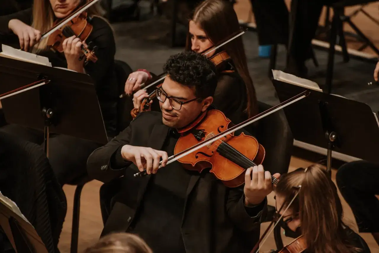 Un homme joue du violon, un instrument à cordes, dans un orchestre. Il est entouré d'autres musiciens.