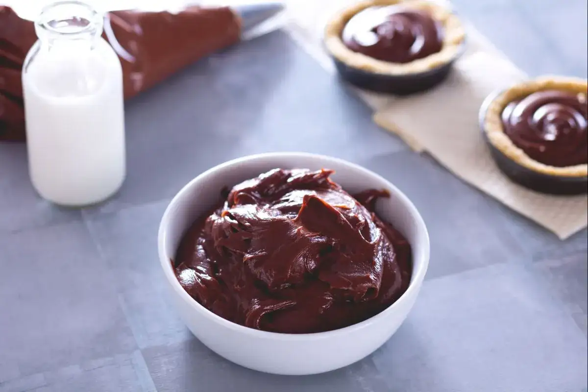 Un bol de ganache chocolat au lait onctueuse, prêt à garnir des tartelettes. Une bouteille de lait et une poche à douille complètent la scène.