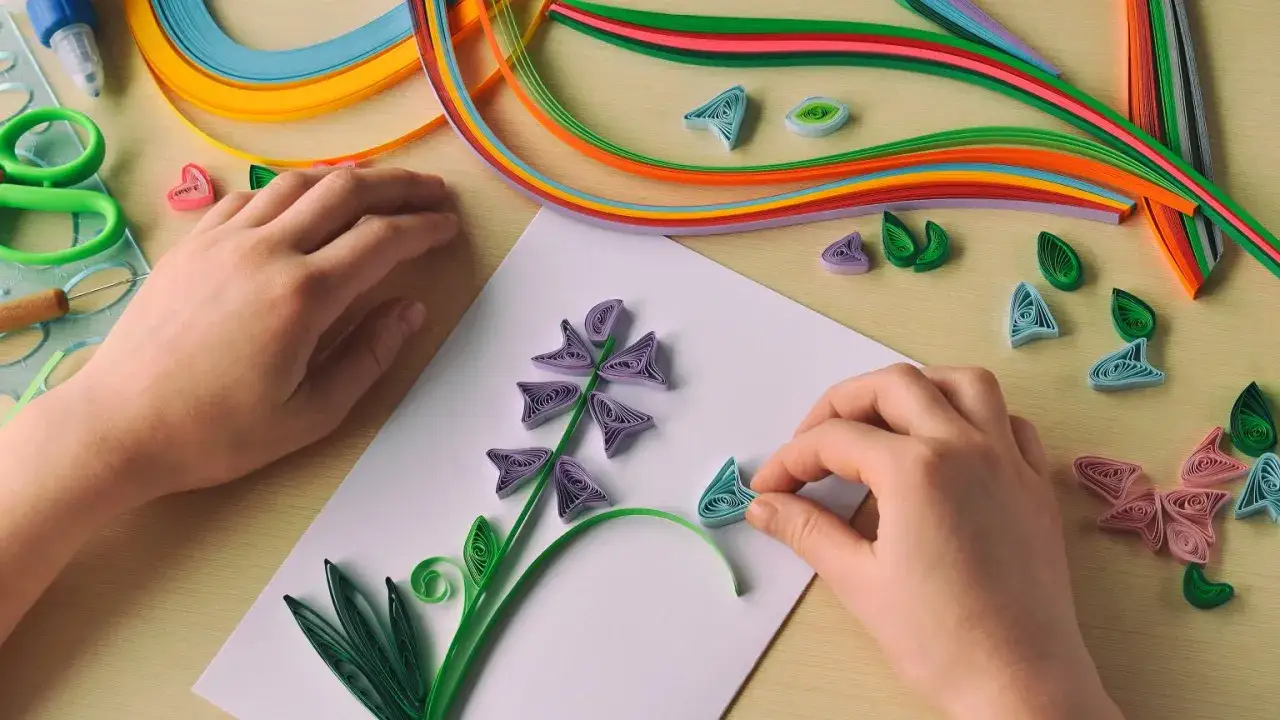 Ręce tworzą kwiat z kolorowych pasków papieru. To właśnie quilling – sztuka tworzenia ozdób z zwiniętych papierowych pasków.