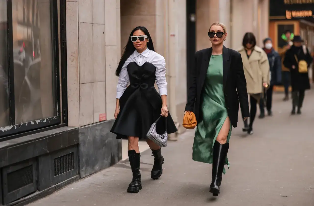 Dos mujeres pasean con un traje con botas. Una lleva un vestido negro con camisa blanca y la otra un vestido verde con blazer negro.