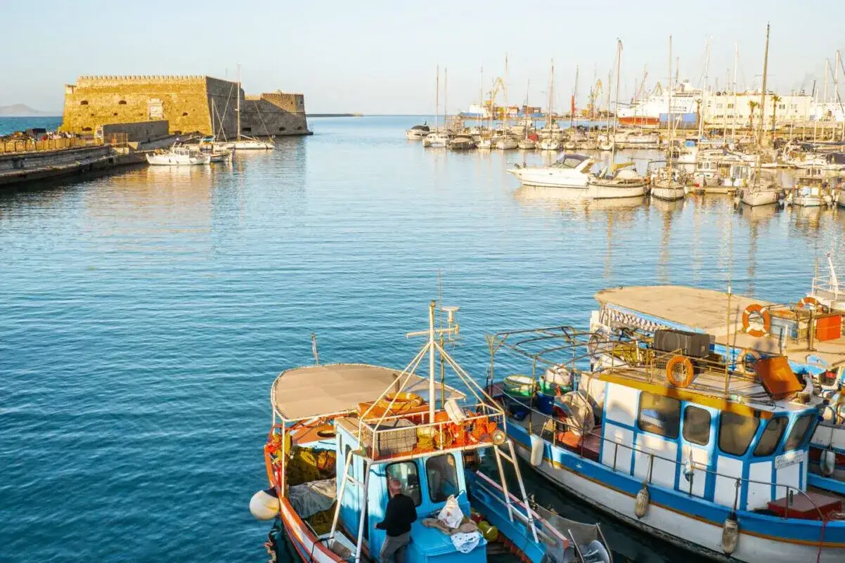 Port w Heraklionie na Krecie. W oddali widać zabytkową fortecę, a na wodzie cumują liczne łodzie i jachty.