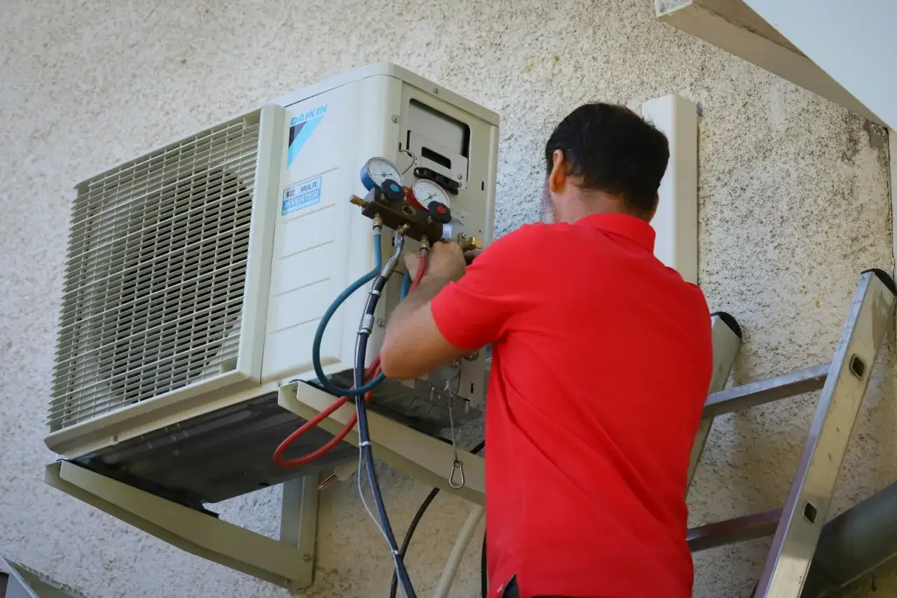 Un technicien en chemise rouge est en train de recharger la clim maison, avec des manomètres connectés à l'unité extérieure.