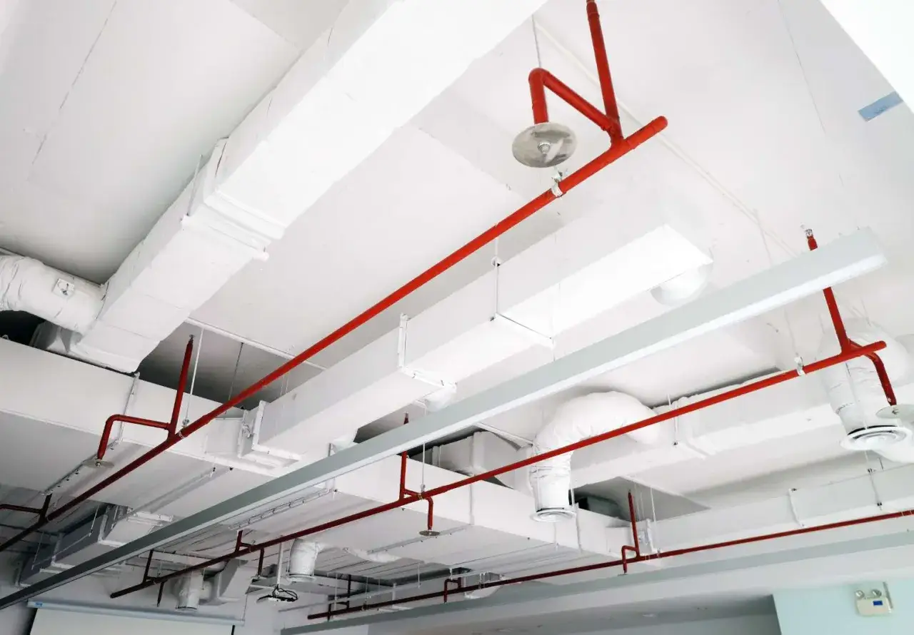 Rote Rohre einer Sprinkleranlage verlaufen über weiße Lüftungskanäle an der Decke.