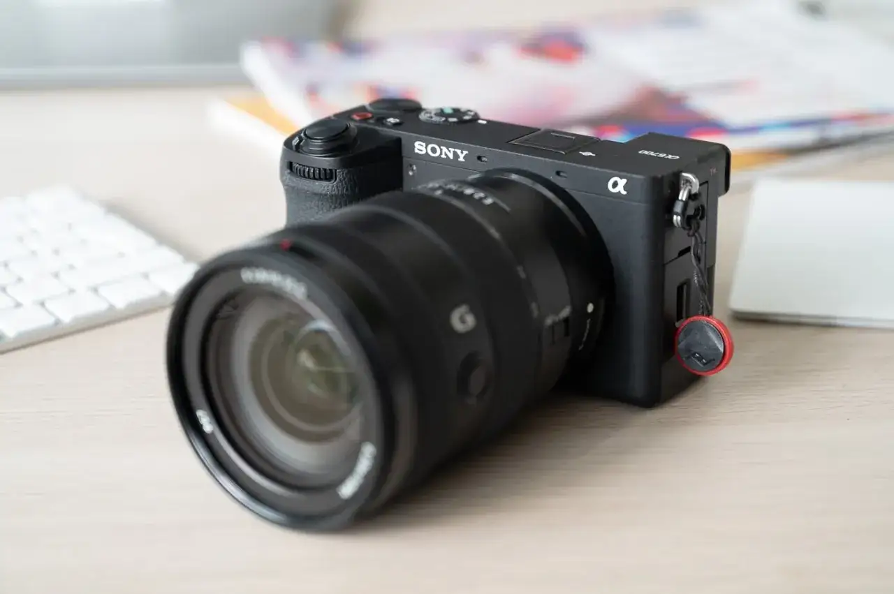 Appareil photo Sony Alpha a6700 avec objectif, pr&ecirc;t pour un test. Un clavier blanc et des documents flous sont en arri&egrave;re-plan.