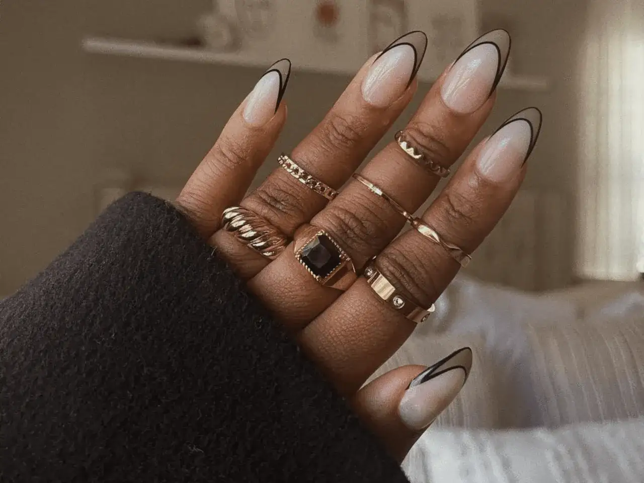 En hand med eleganta press on nails i fransk manikyr med svart kant. Flera guldringar pryder fingrarna.