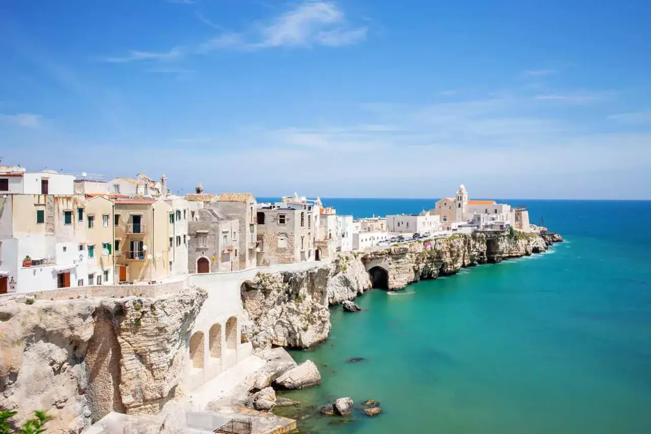 Vacker kuststad i **Puglia, Italien**, med vita hus som kl&auml;ttrar uppf&ouml;r klipporna mot det turkosbl&aring; havet.
