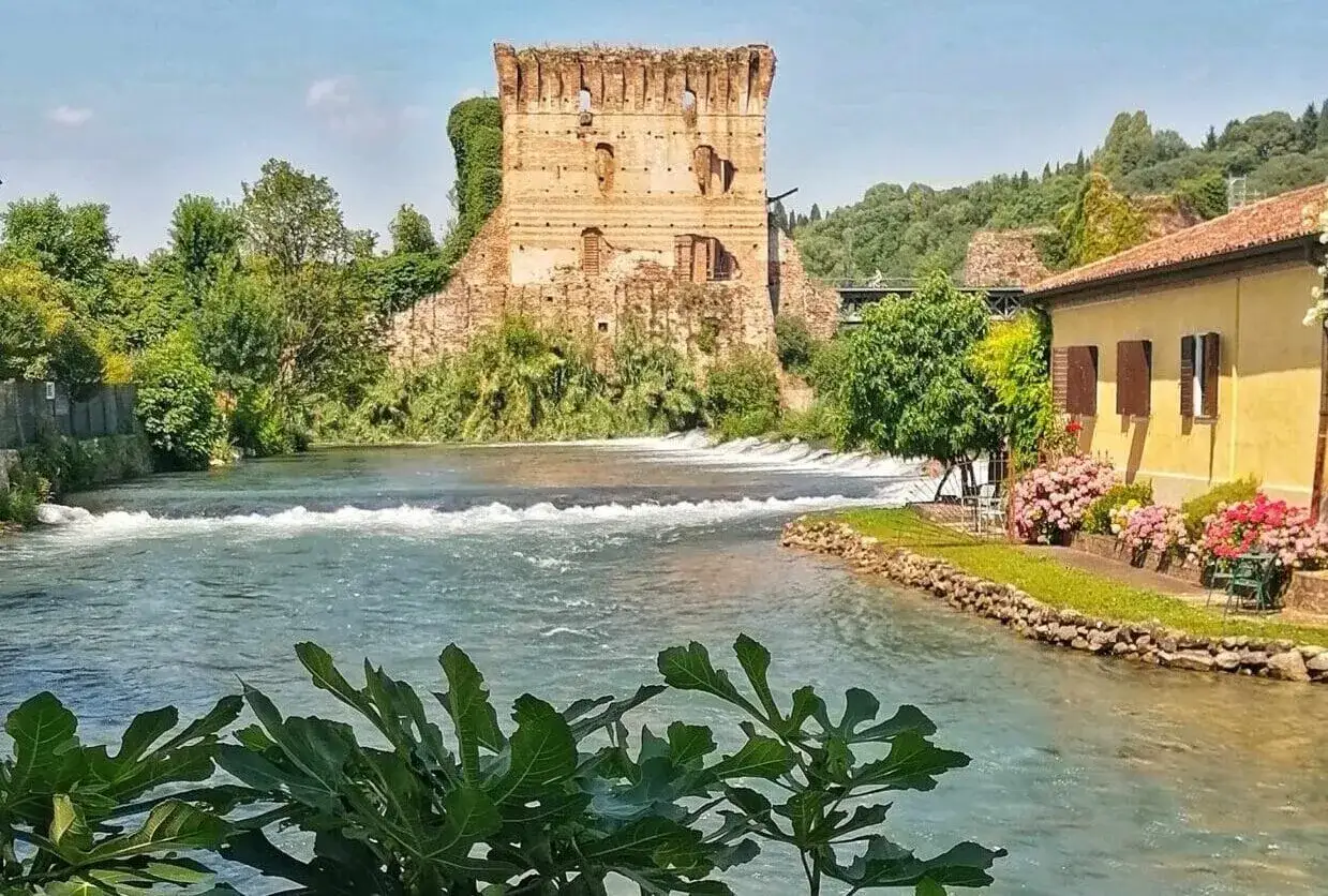 Il fiume Mincio scorre placido vicino a un antico borgo, con una torre in rovina e una casa gialla fiorita.