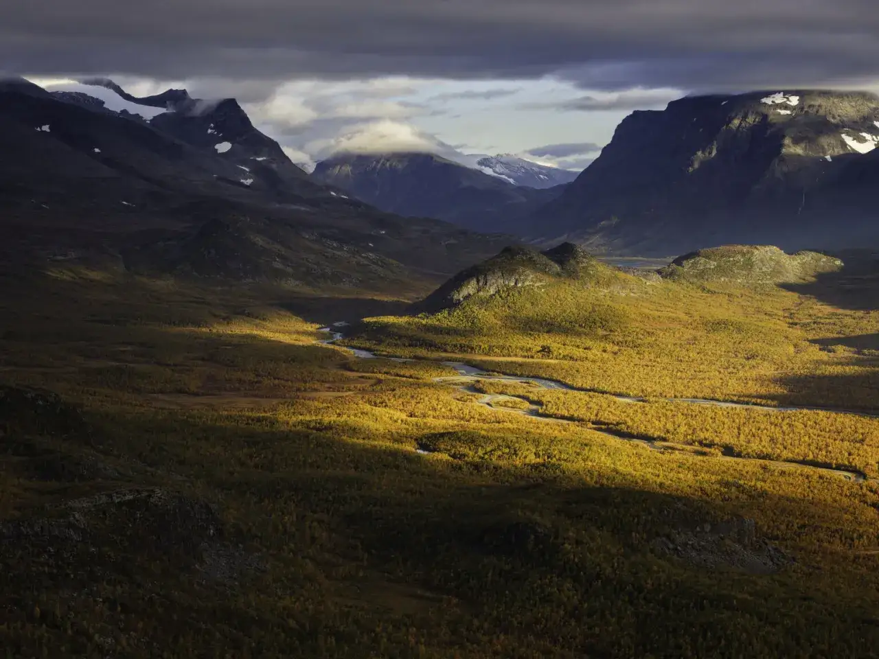 Magnus Lindbom f&aring;ngar h&ouml;stens gyllene ljus &ouml;ver fj&auml;ll och en slingrande &auml;lv. Dramatiska bergskedjor reser sig mot en molnig himmel.
