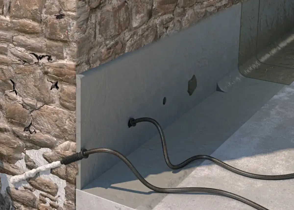 Mauerwerk Injektion selber machen: Ein Schlauch führt in ein gebohrtes Loch in einer Betonwand, während daneben eine Steinwand mit Rissen und einer Füllung zu sehen ist.
