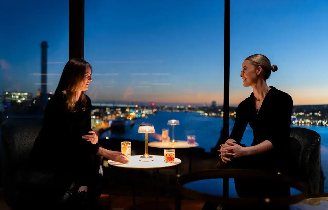 Två kvinnor samtalar vid ett bord på en **rooftop bar i Göteborg** med utsikt över stadens ljus vid skymningen.