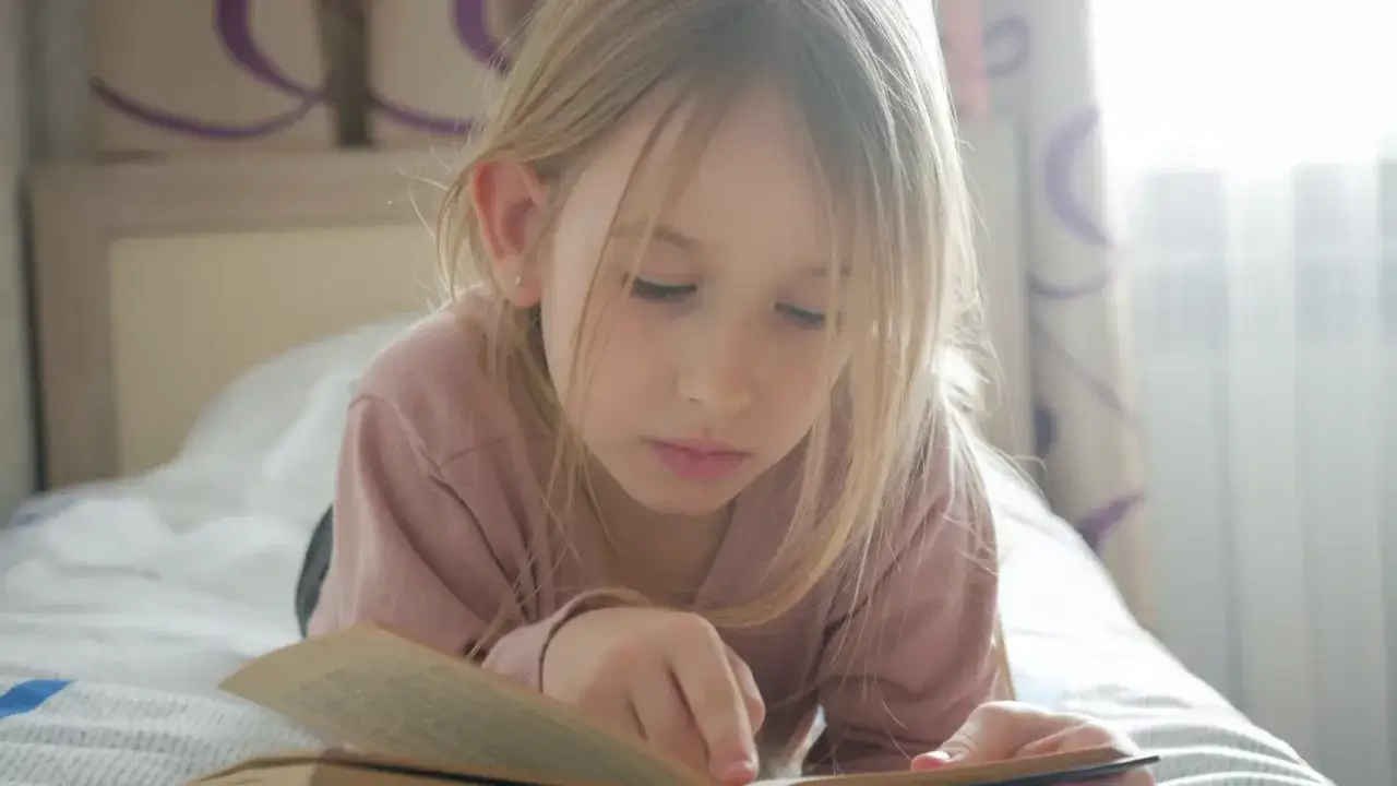 Dziewczynka z blond włosami leży na łóżku i czyta książkę. Idealne książki dla 12 latki, które wciągną ją w świat przygód.