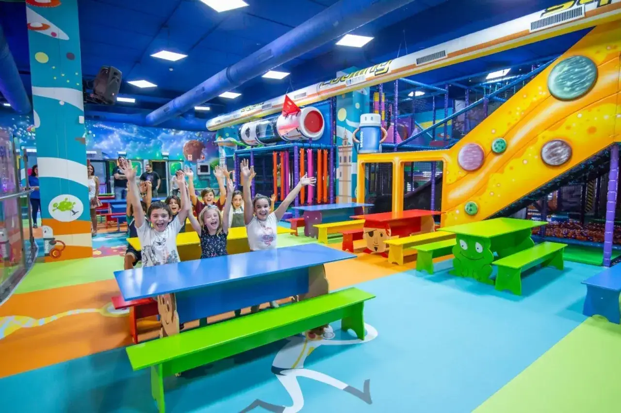 Niños felices saludan desde un centro comercial con juegos para niños, con toboganes, túneles y mesas coloridas.