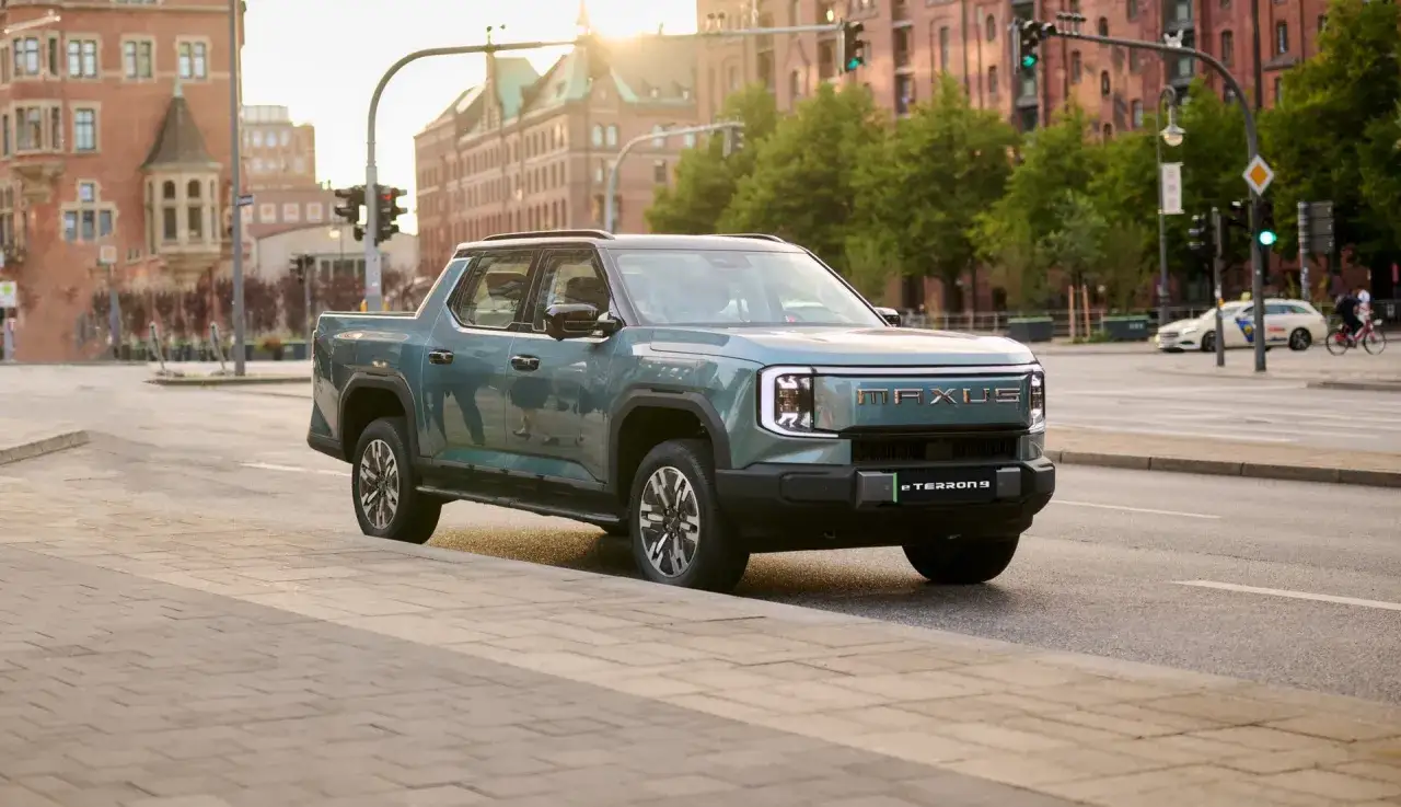 En gr&ouml;n el pickup, Maxus eTerron 9, st&aring;r vid en trottoar i en stadsmilj&ouml; med tegelbyggnader och tr&auml;d i bakgrunden.