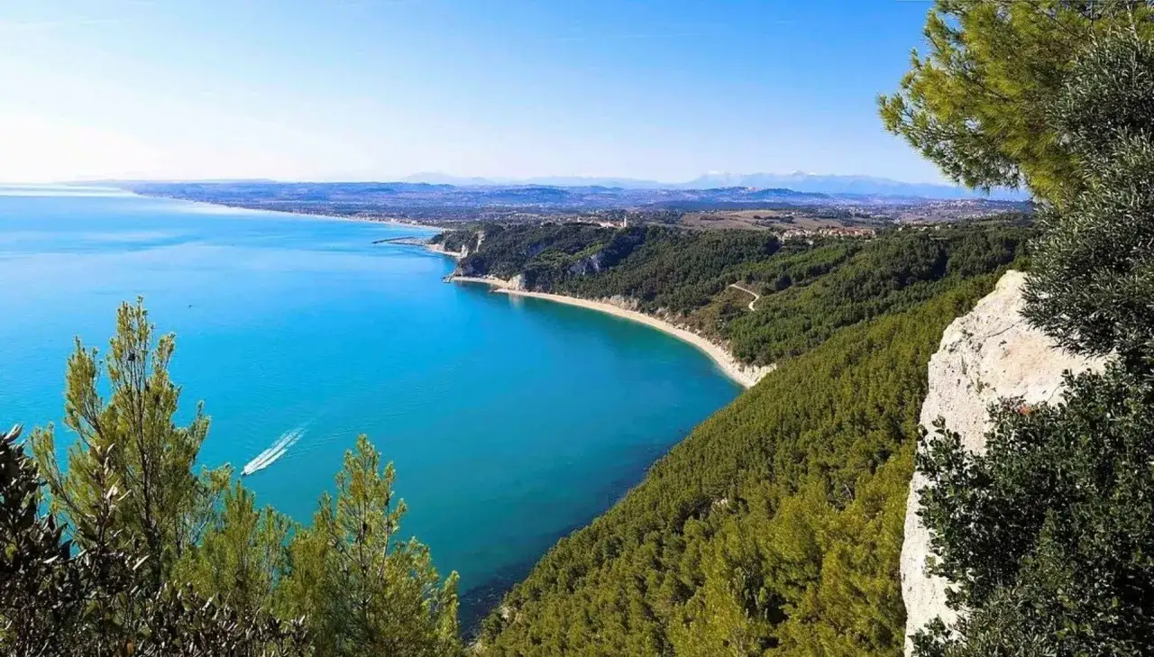 Vista mozzafiato della Riviera del Conero: mare azzurro, spiagge dorate e pineta rigogliosa. Una barca solca le acque cristalline.