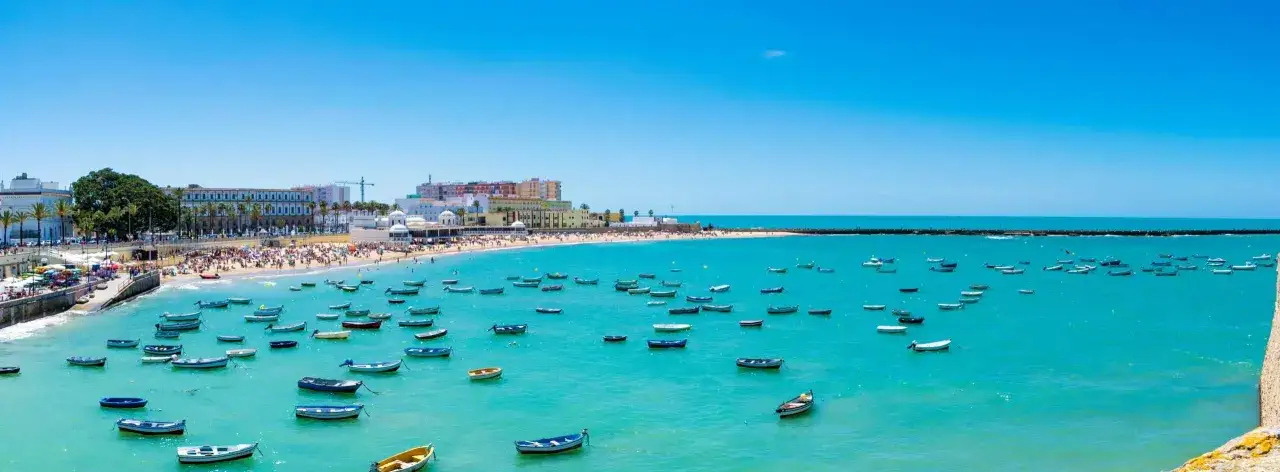 Bunte Boote auf dem t&uuml;rkisfarbenen Wasser vor dem belebten **Cadiz Strand** mit der Stadtkulisse im Hintergrund.