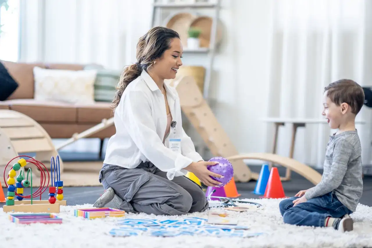 Ergotherapie bei ADHS: Eine Therapeutin spielt mit einem Kind auf einem Teppich, um Konzentration und Feinmotorik zu fördern. Bunte Spielsachen liegen herum.