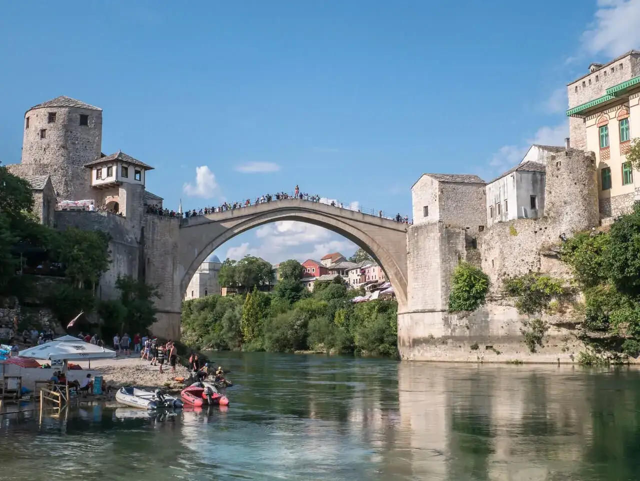 Mostar, Bośnia i Hercegowina: Słynny Stary Most z kamienia, tłumy ludzi na moście i brzegu, łodzie na rzece Neretwie.