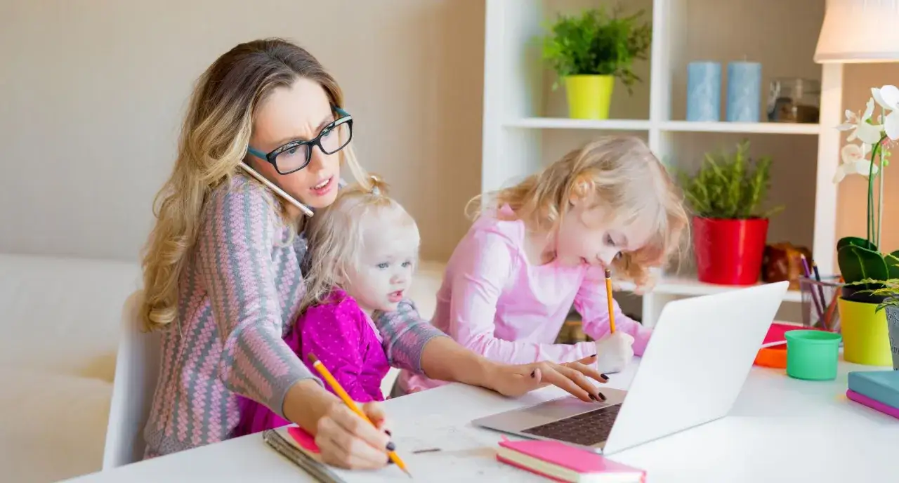 Zabiegana mama z dwójką dzieci, młodsze na kolanach, starsze przy laptopie. Opieka na dziecko do ilu lat? Tu widać, że nawet przy dwójce maluchów.