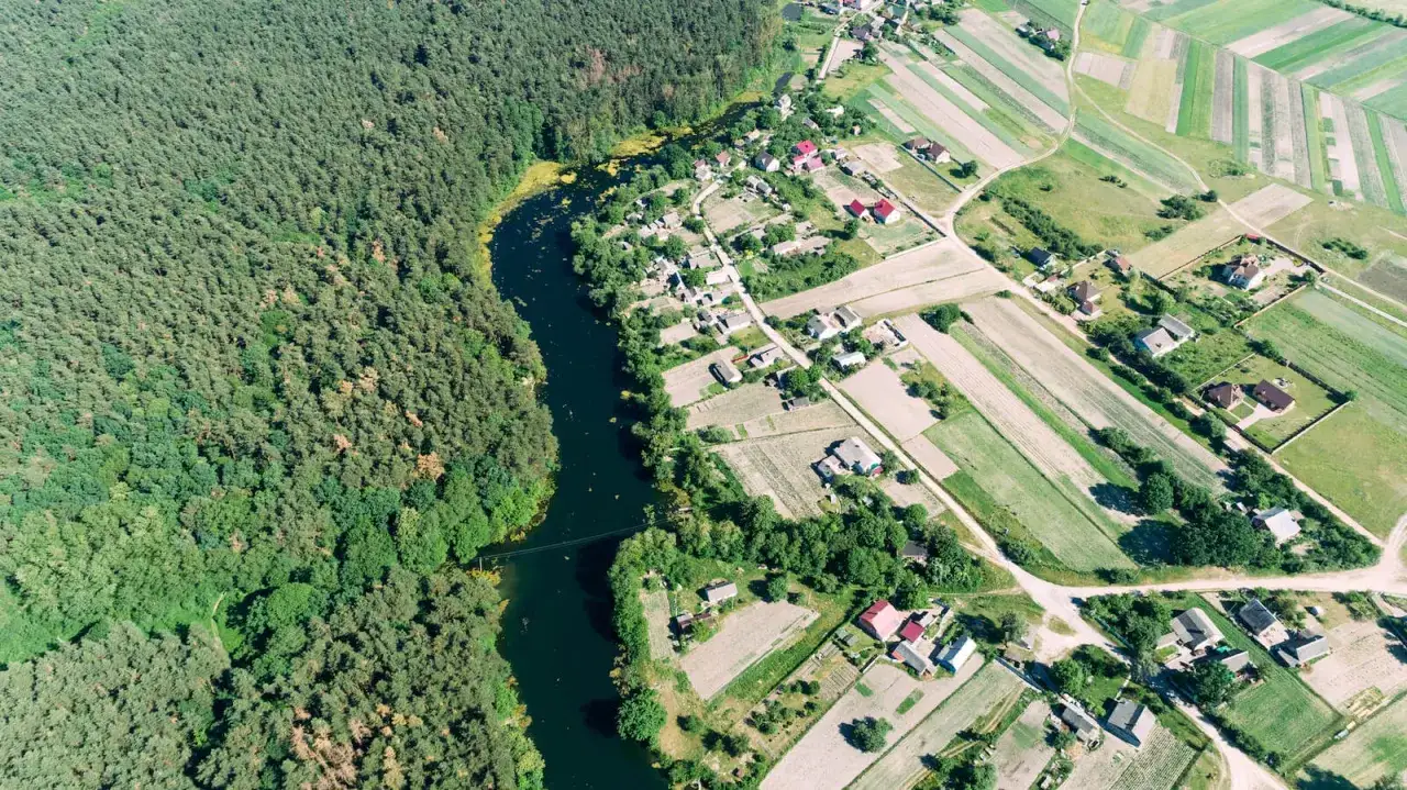 Widok z lotu ptaka na wieś z lasem i ciemną wodą. Widać pola uprawne, domy z czerwonymi dachami i wijący się rów melioracyjny.