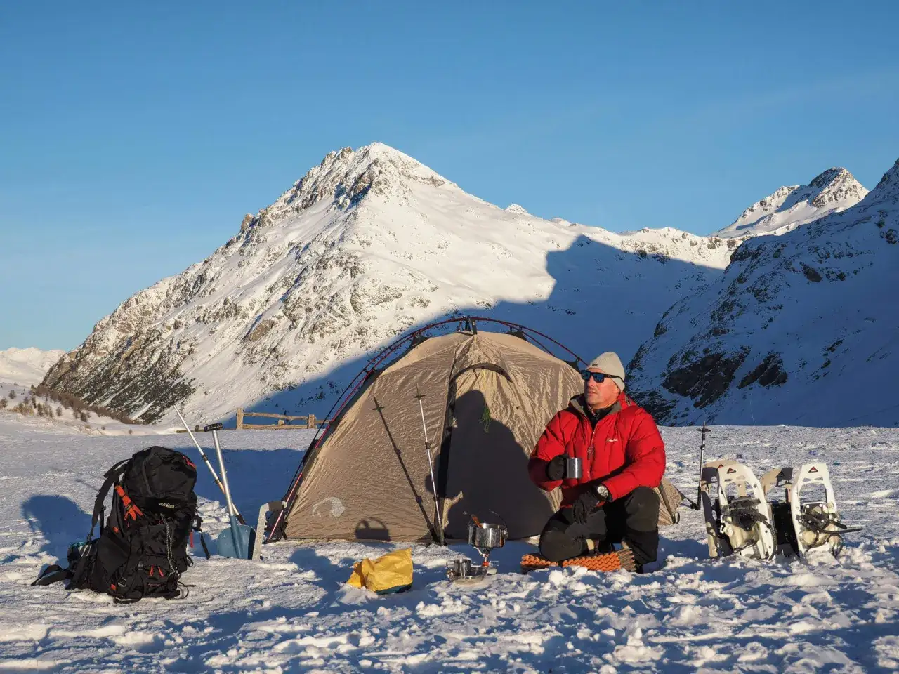 Mann genie&szlig;t hei&szlig;en Tee beim Zelten im Winter vor schneebedeckten Bergen. Schneeschuhe und Rucksack liegen bereit.