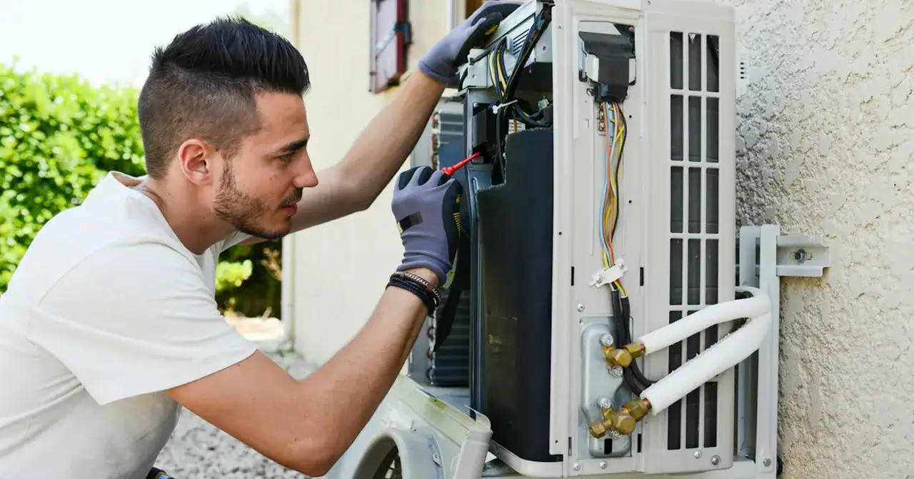 Un technicien vérifie un climatiseur extérieur. Il est concentré sur les fils et les composants. Le **tarif dépannage climatisation** est une préoccupation pour les propriétaires.