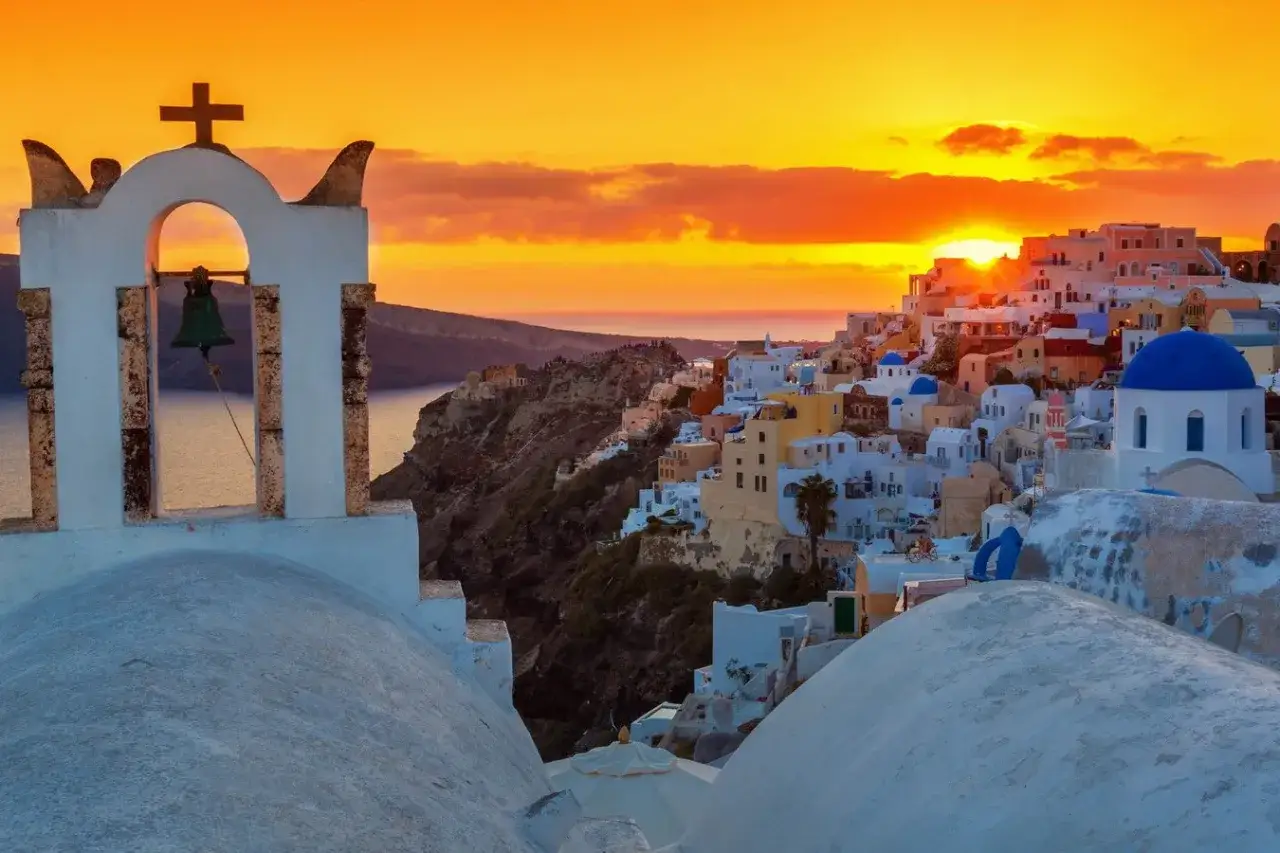 Zachód słońca nad Santorini. Dzwonnica z krzyżem i dzwonem na pierwszym planie, a w tle białe domy z niebieskimi kopułami. Idealne miejsce na wycieczkę z krety.