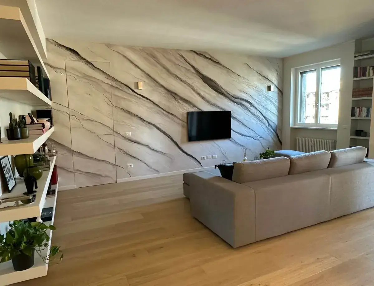 Soggiorno moderno con parete effetto marmo, un divano beige e libreria. Lo **stucco materico** crea un'atmosfera elegante e unica.