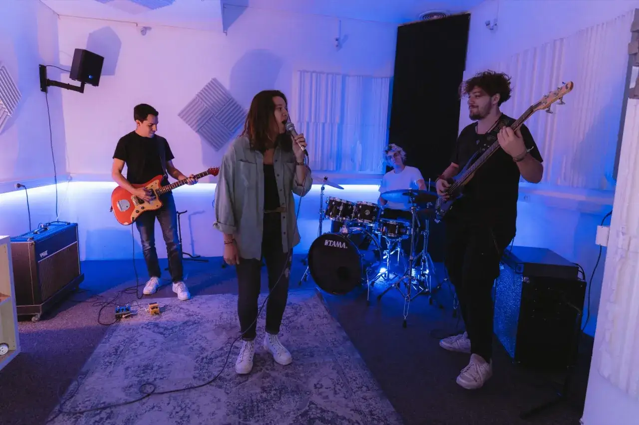 Un groupe de musique répète dans un studio. La chanteuse est au micro, le guitariste joue, le bassiste est prêt à rejoindre le groupe.